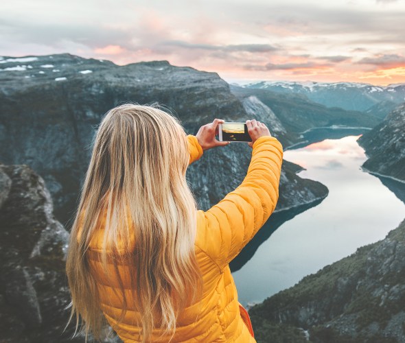 A woman in a yellow jacket takes a picture of a fjord in Norway at sunrise or sunset