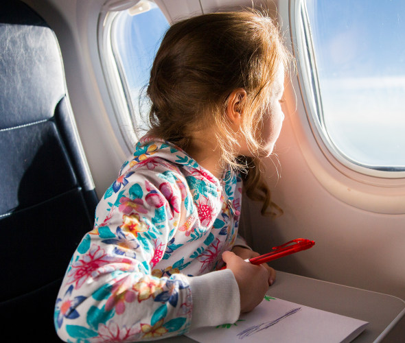 A little girl takes a break from coloring to look out the airplane window.