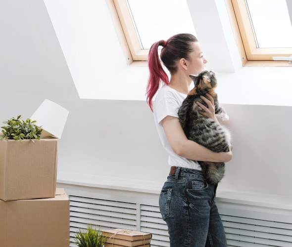 A woman holds a cat in front of stacked moving boxes.