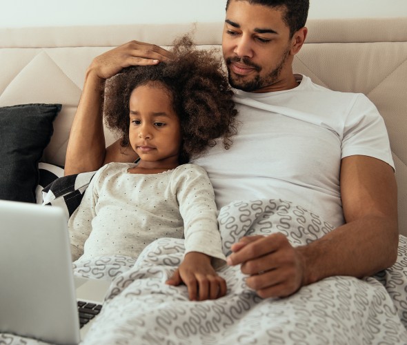 A father and daughter watch tv on a computer in bed.