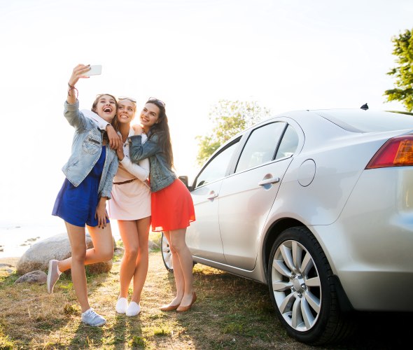 Three young teen girls smile for a selfie next to the drivers car