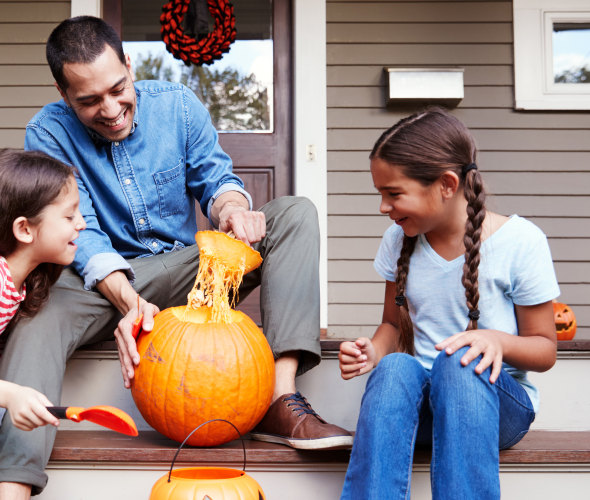 A dad carves a pumpkin with his daughters on their porch.