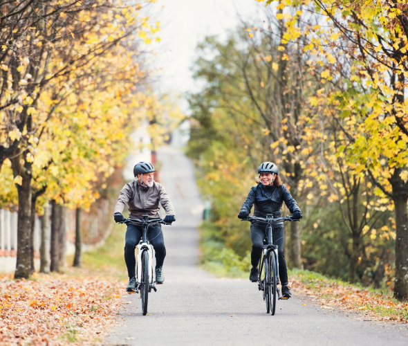A couple rides their e-bikes on a tree-lined bike path.
