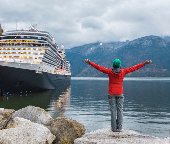 A woman stands on the shore with her Alaskan cruise ship in front of her.