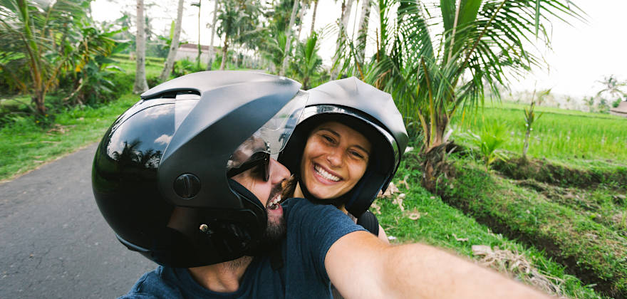 Couple takes a selfie while riding a scooter through a tropical locale