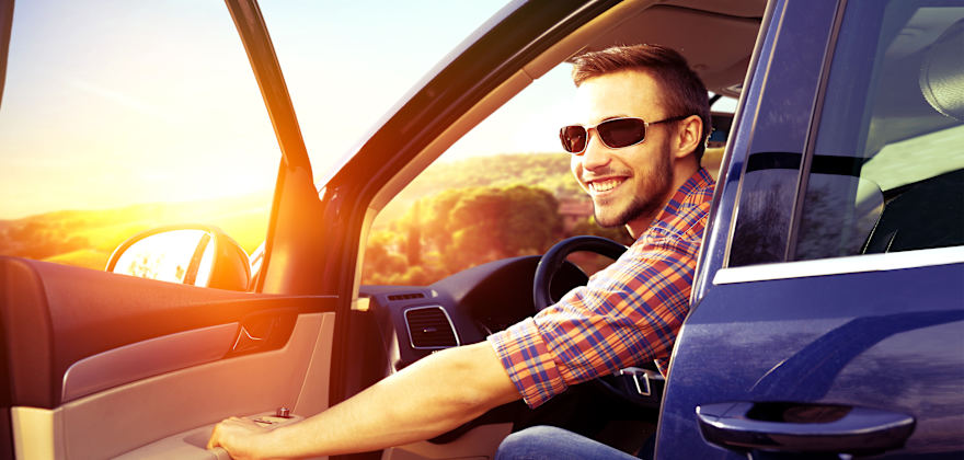 A man smiling sits in his blue car holding the door open with the sun shining brightly on the horizon