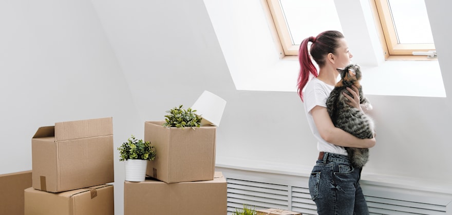 A woman holds a cat in front of stacked moving boxes.