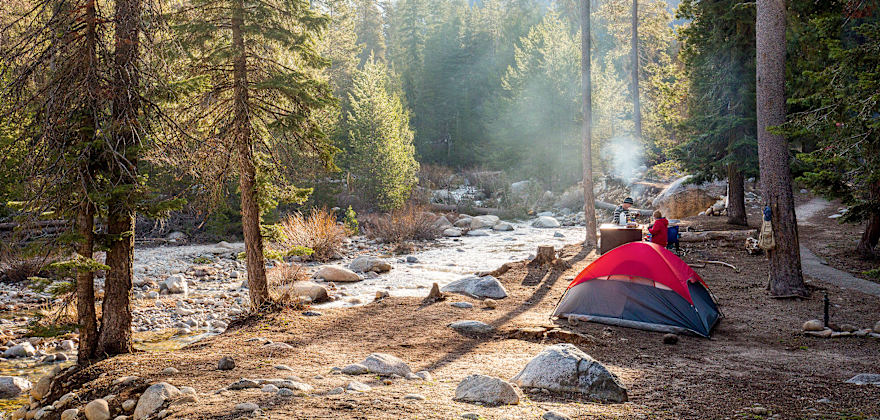 pair of campers with tent pitched beside stream are dressed for cool temps on sunny day at Dorst Creek Campground in Sequoia National Park, California