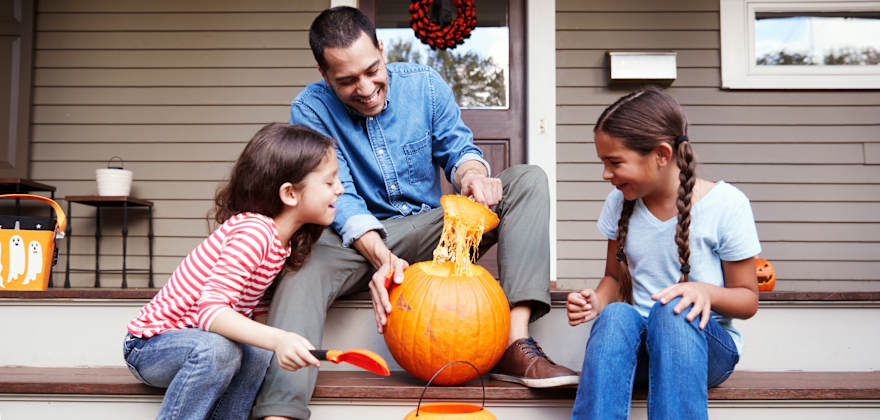 A dad carves a pumpkin with his daughters on their porch.