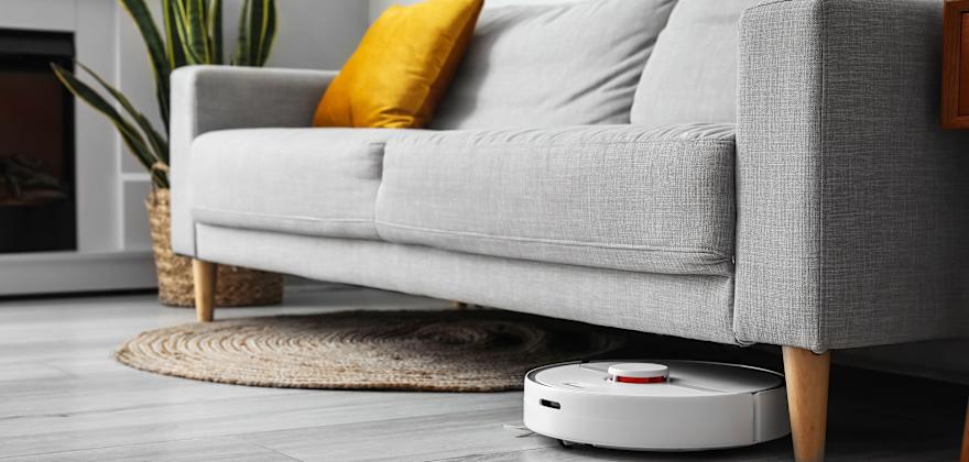 A smart robot vacuum cleans under a gray sofa.