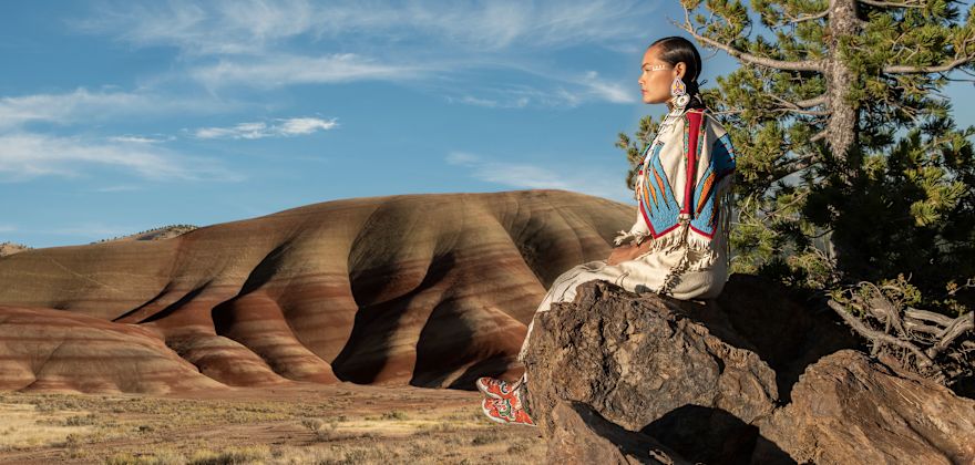 Acosia M. Red Elk in a buckskin dress sitting on a rock overlooking the Oregon landscape.