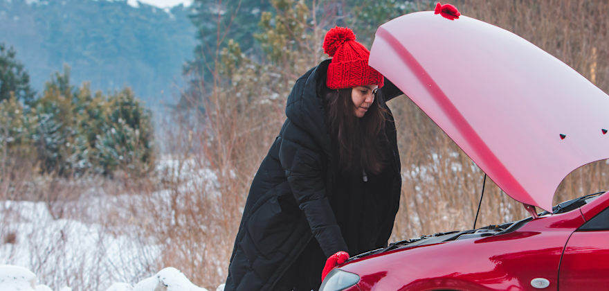 A woman in a coat and hat looks under the hood of her red car.