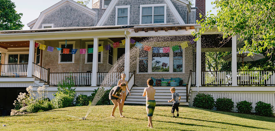Kids play in a sprinkler in front of a wood-shingled house.