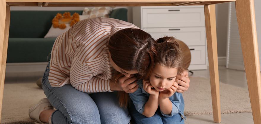 A mom and her daughter drop, cover, and hold on under a table during an earthquake.