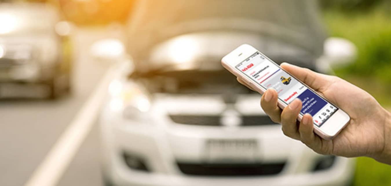 Hand of a AAA Member using the AAA app on their cellphone to call for roadside assistance, with broken down car in the background.