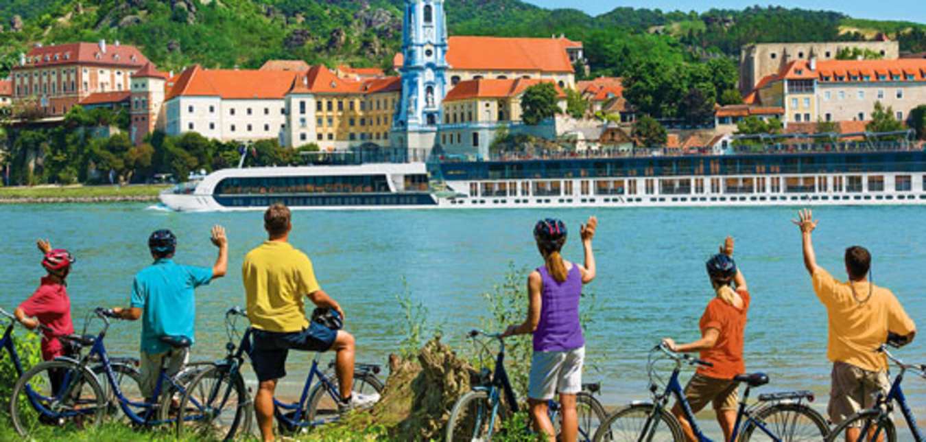 group of bicyclists on a river bank waving to amawaterways ship