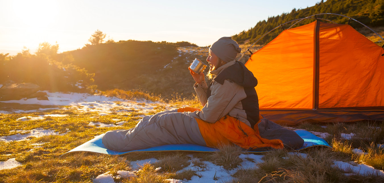 A camper sits on a sleeping pad and sleeping bag on frozen ground while drinking a coffee.