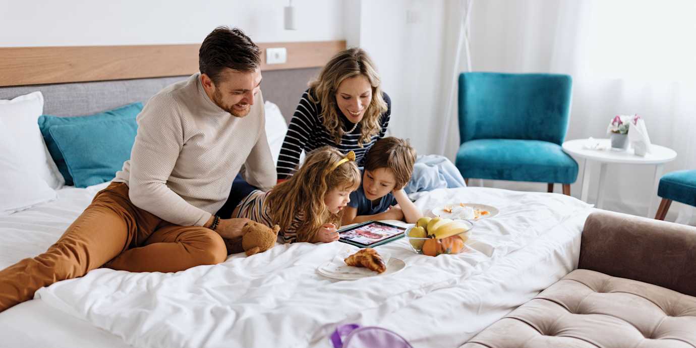 A family watches something on a tablet on a hotel bed.