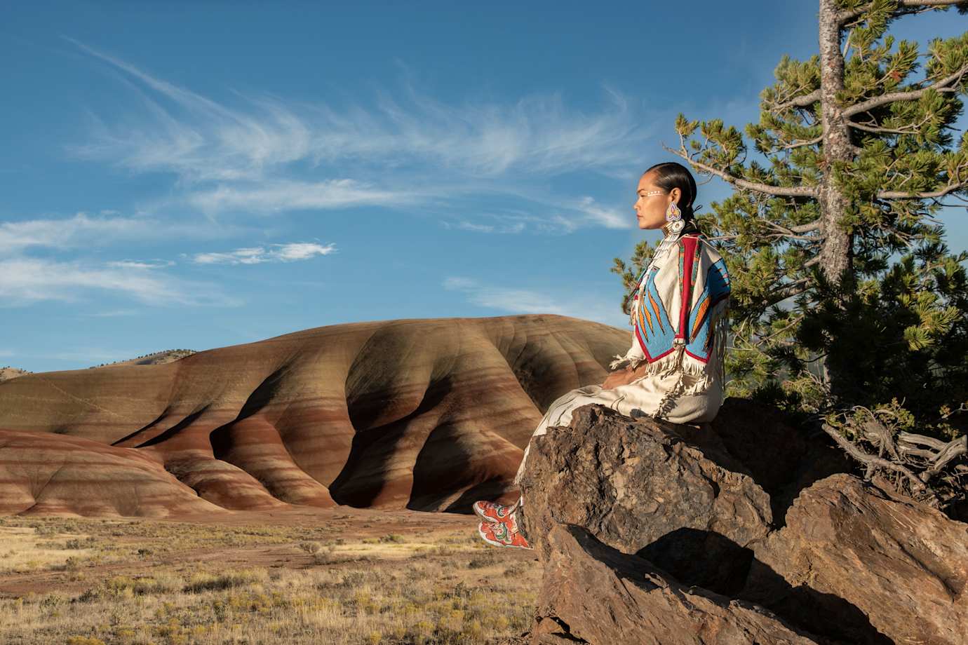 Acosia M. Red Elk in a buckskin dress sitting on a rock overlooking the Oregon landscape.