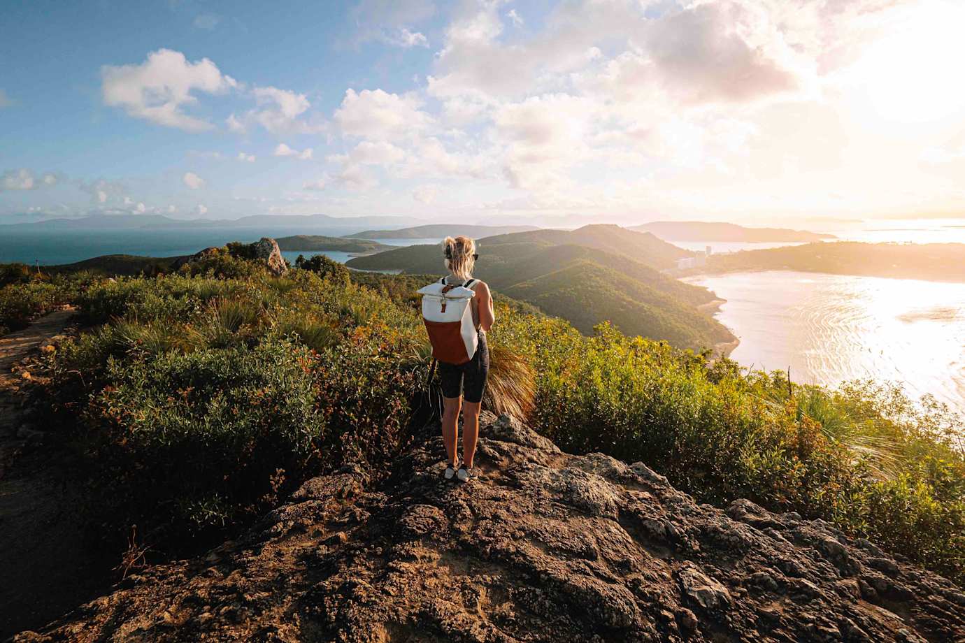a woman wearing shorts and a backpack stands on top of a mountain looking out at the ocean in the sun