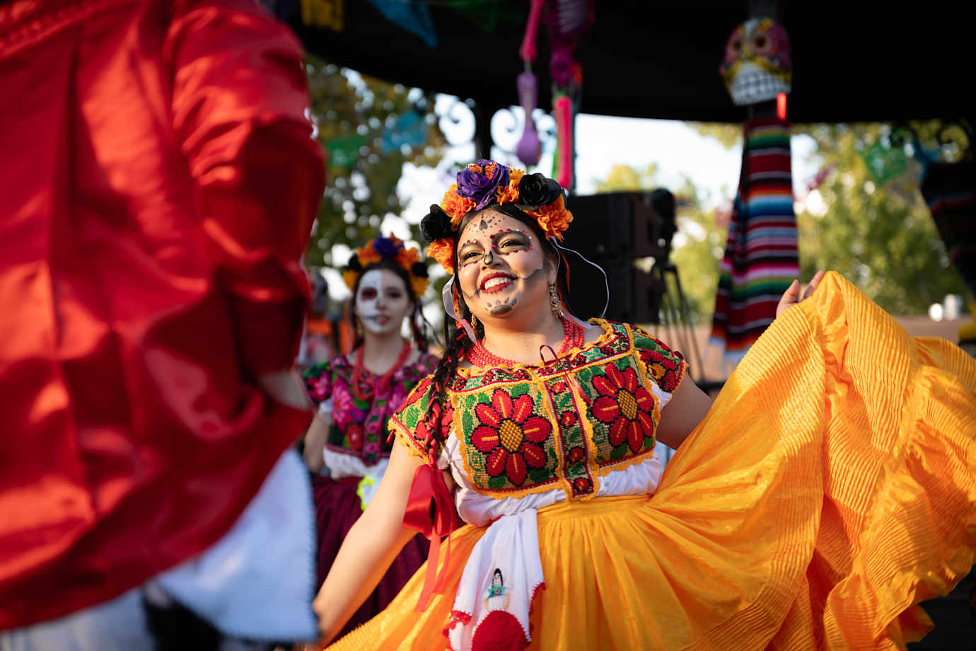 a woman dances in a colorful dress and face paint at a Day of the Dead celebration.