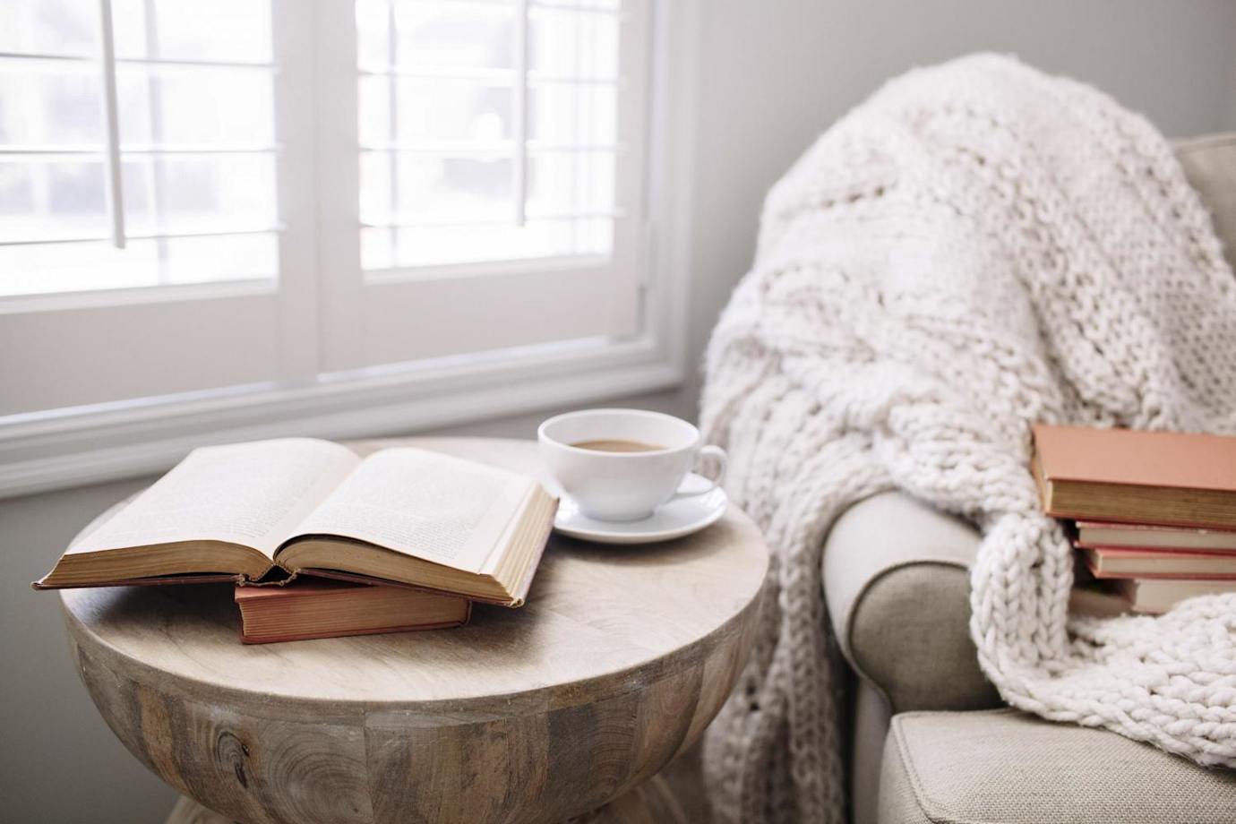 cozy armchair by window with throw, side table, books, cup of tea.