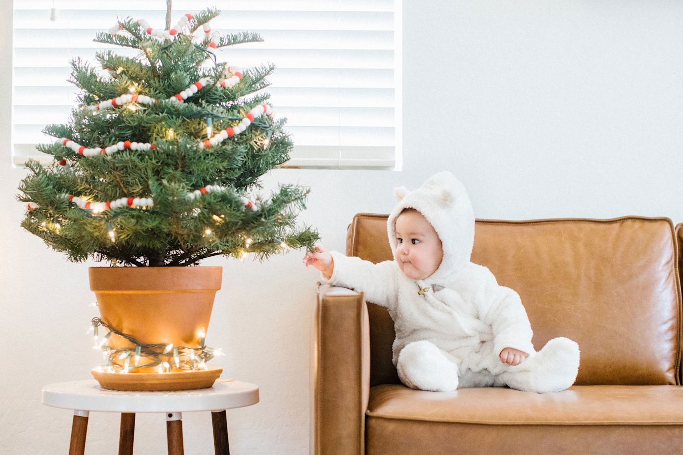 A baby on a leather couch reaches out to touch a tabletop Christmas tree.