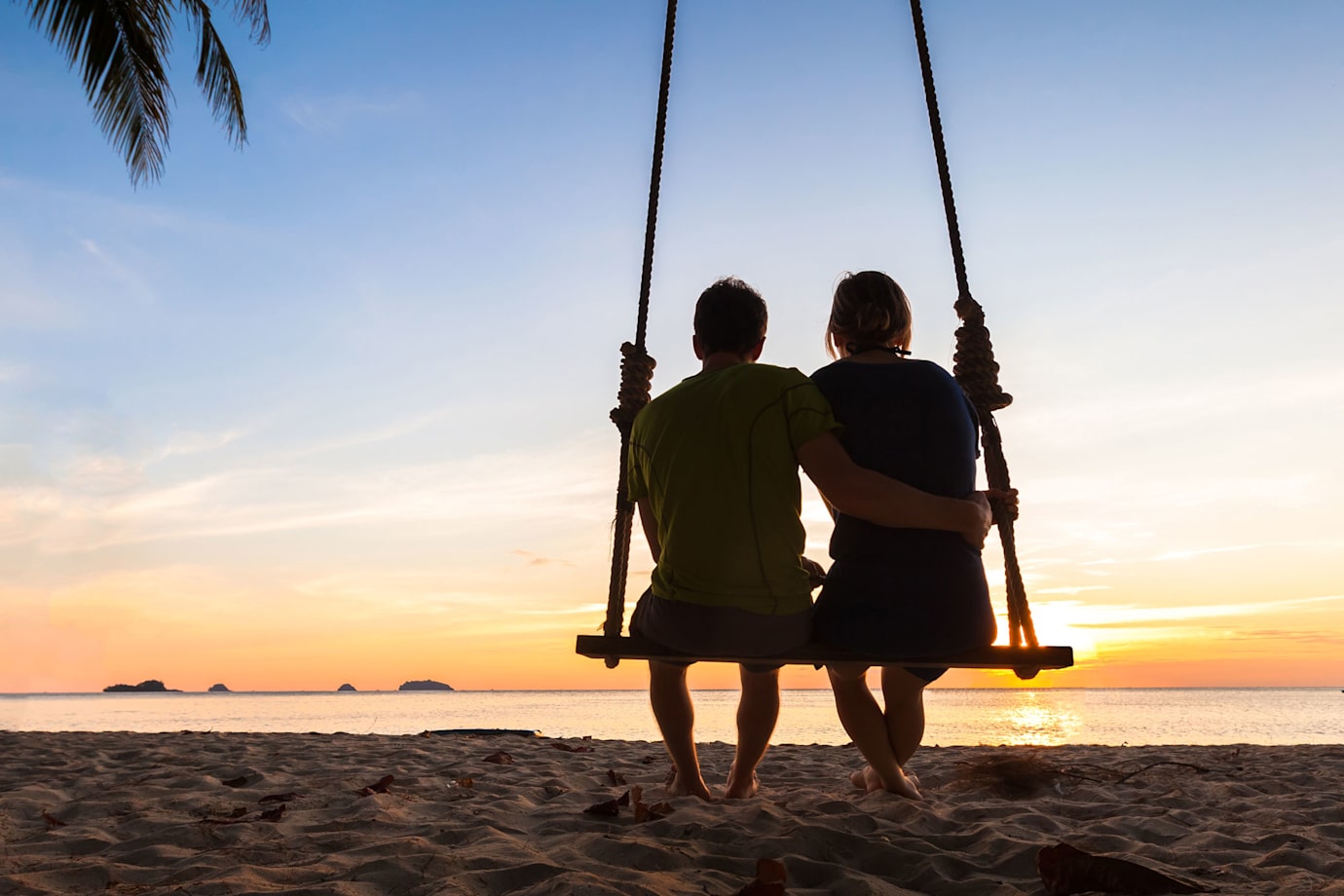 newlyweds sit on a swing together to watch a tropical sunset