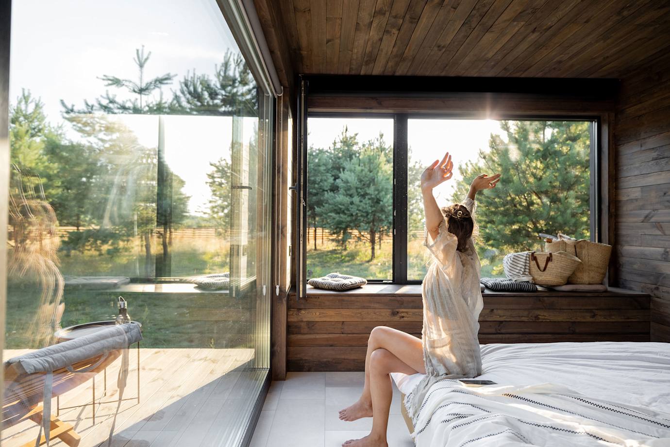 A woman stretches at the foot of her bed as morning light streams into her hotel room.