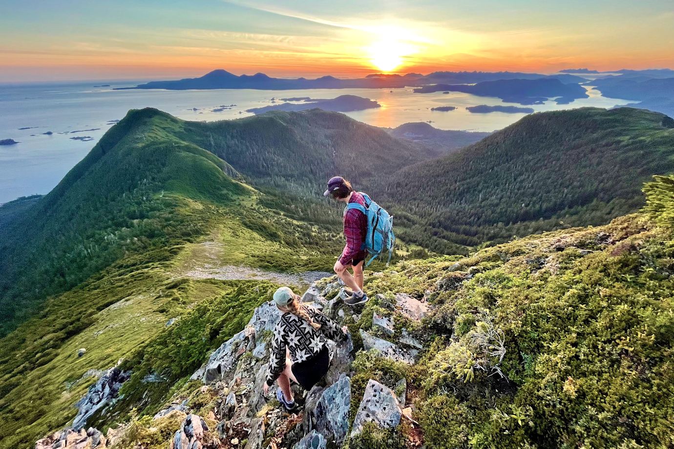Two hikers scramble on rocks along trail overlooking Sitka sound at sunset.