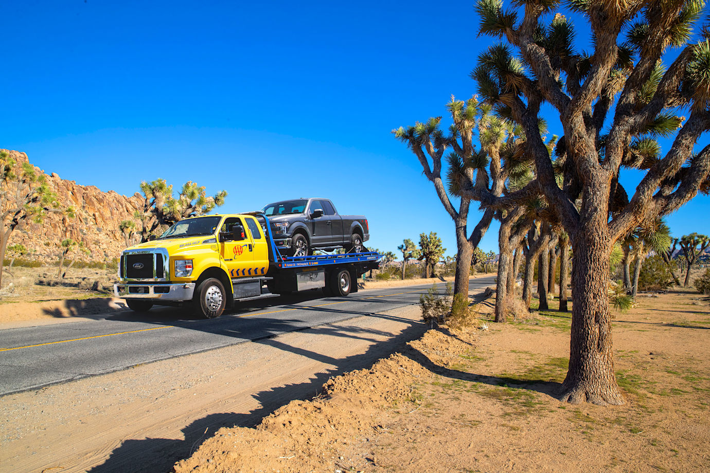 A AAA tow truck drives through Joshua Tree with a pickup on the back.