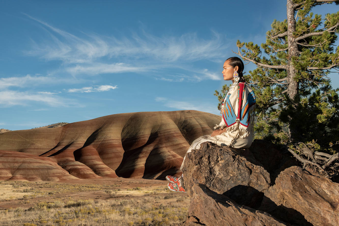 Acosia M. Red Elk in a buckskin dress sitting on a rock overlooking the Oregon landscape.