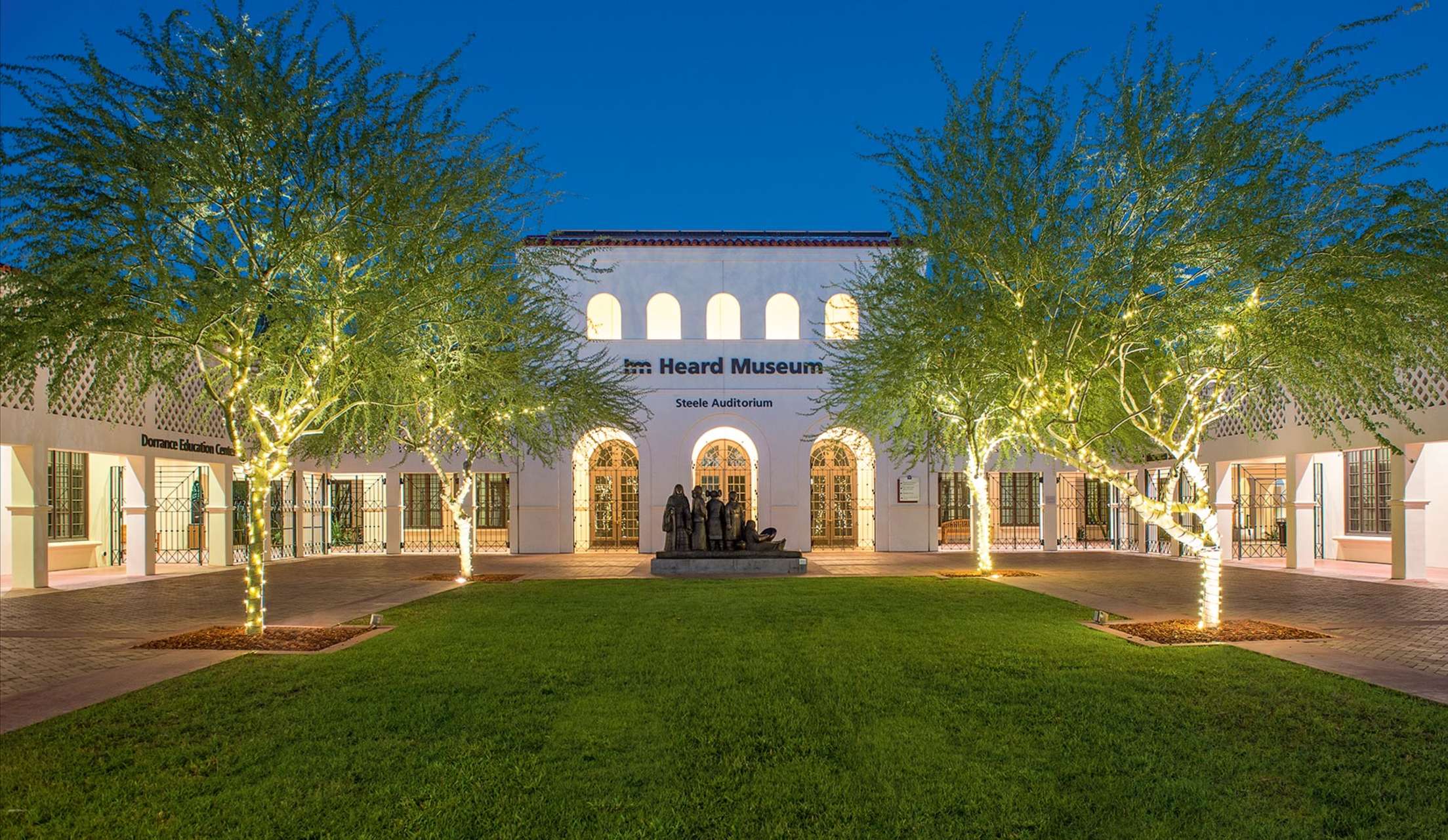 Heard Museum entrance across courtyard with figural sculpture and lawn lined by cottonwood trees with string lights, at twilight in Phoneix, Arizona, picture