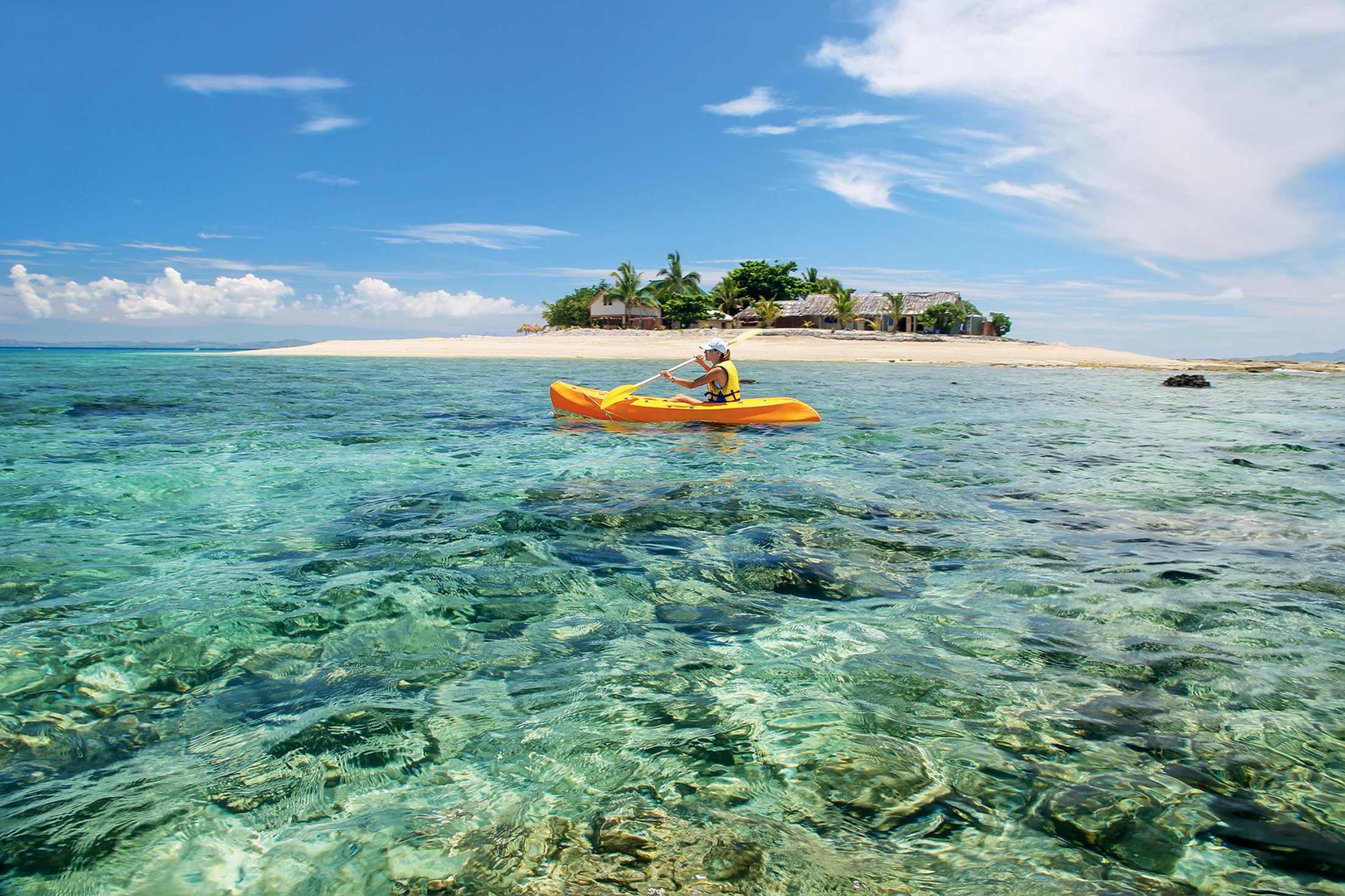 a solo kayaker paddles through translucent waters passing a tiny island in the Mamanucas Islands, part of Fiji
