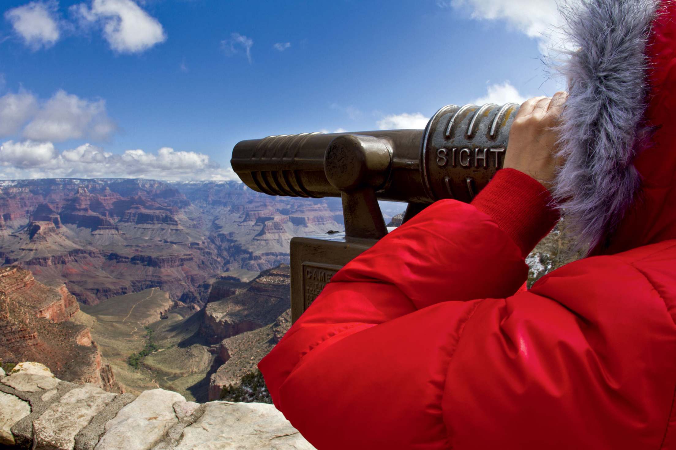 viewer in red winter parka with scope checks out the rock formations from the Rim Trail at Grand Canyon National Park in Arizona