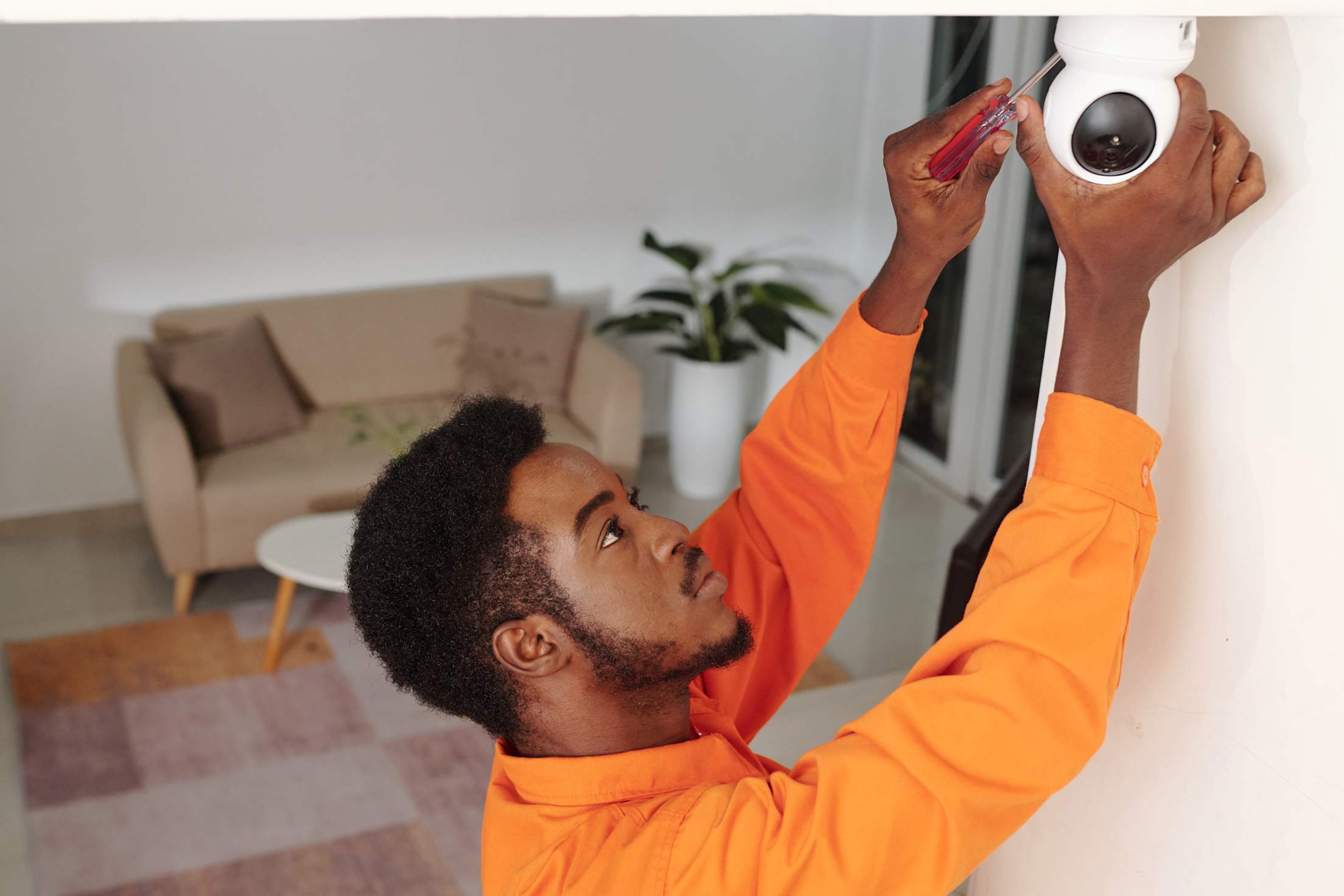 A man in an orange shirt installs a new indoor security camera on the ceiling.