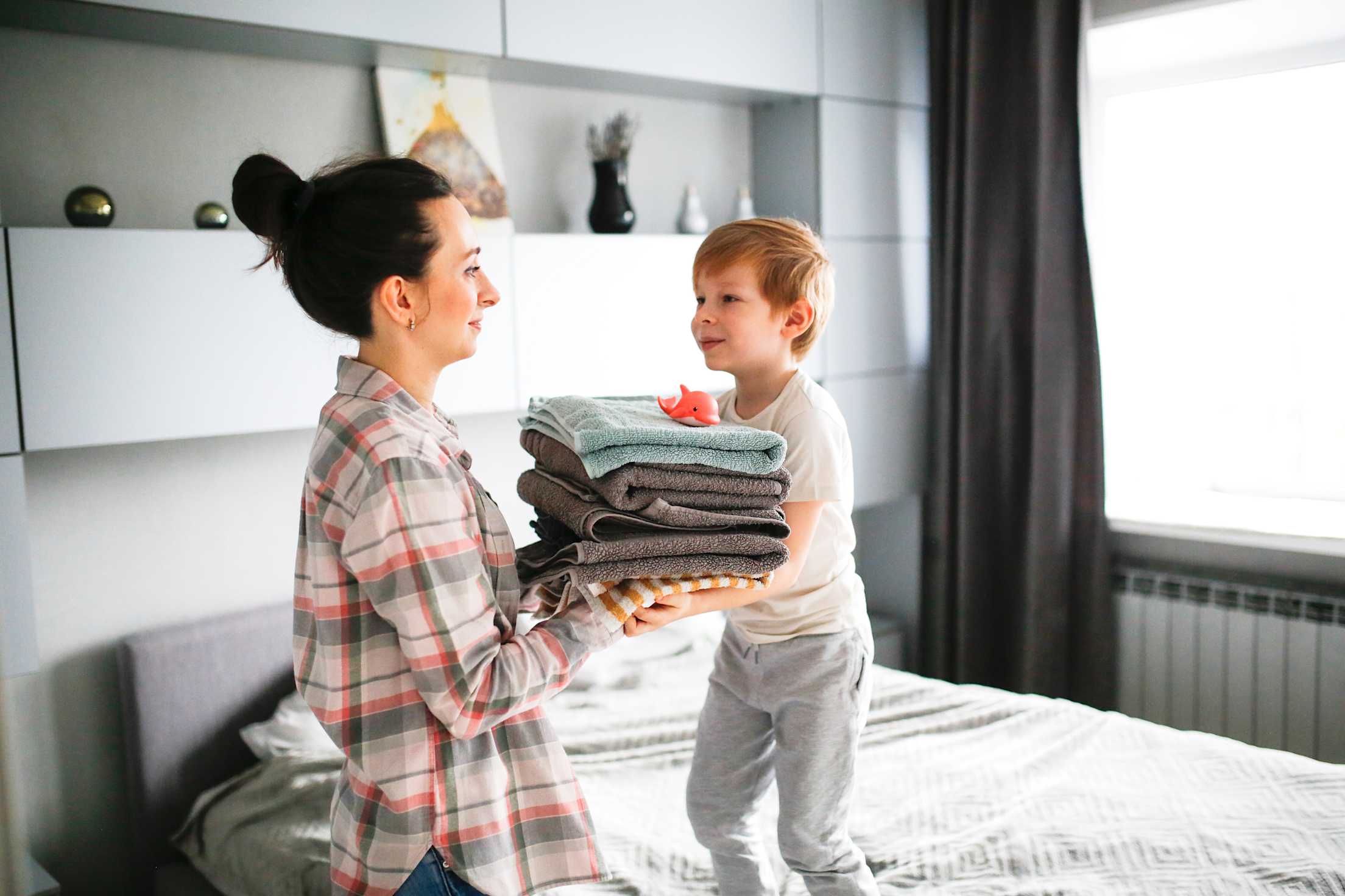 A mom hands her son a stack of towels with a bath toy on top.