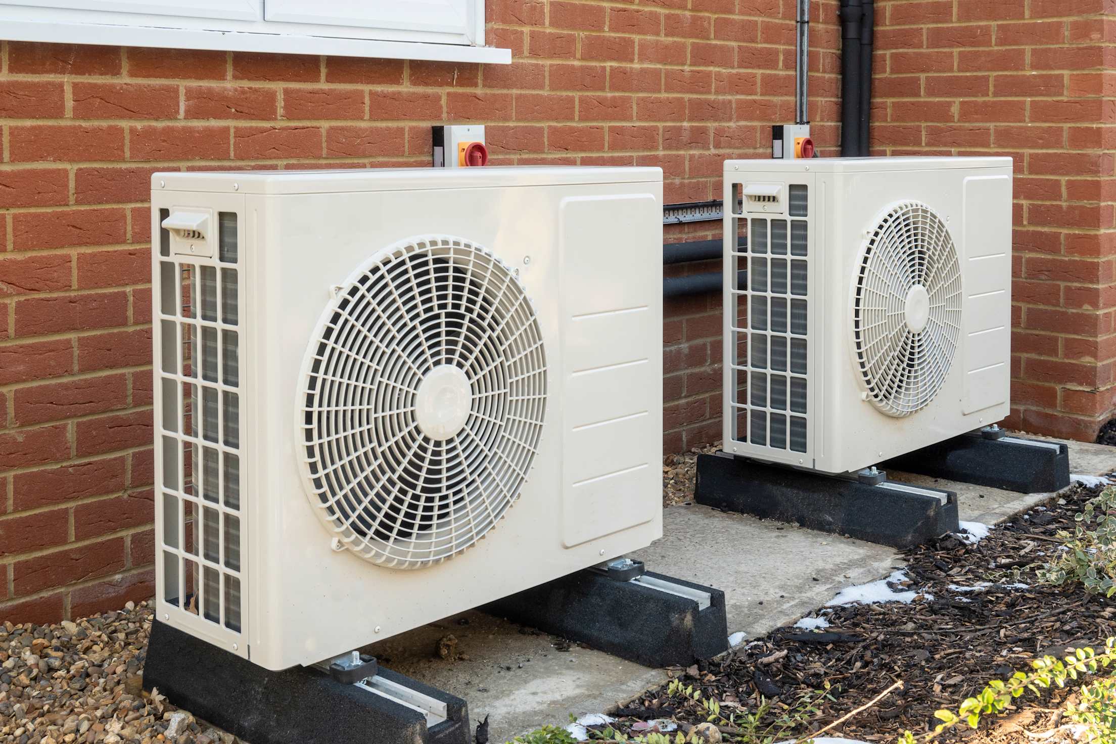 Two external air heat pump units outside of a brick home.