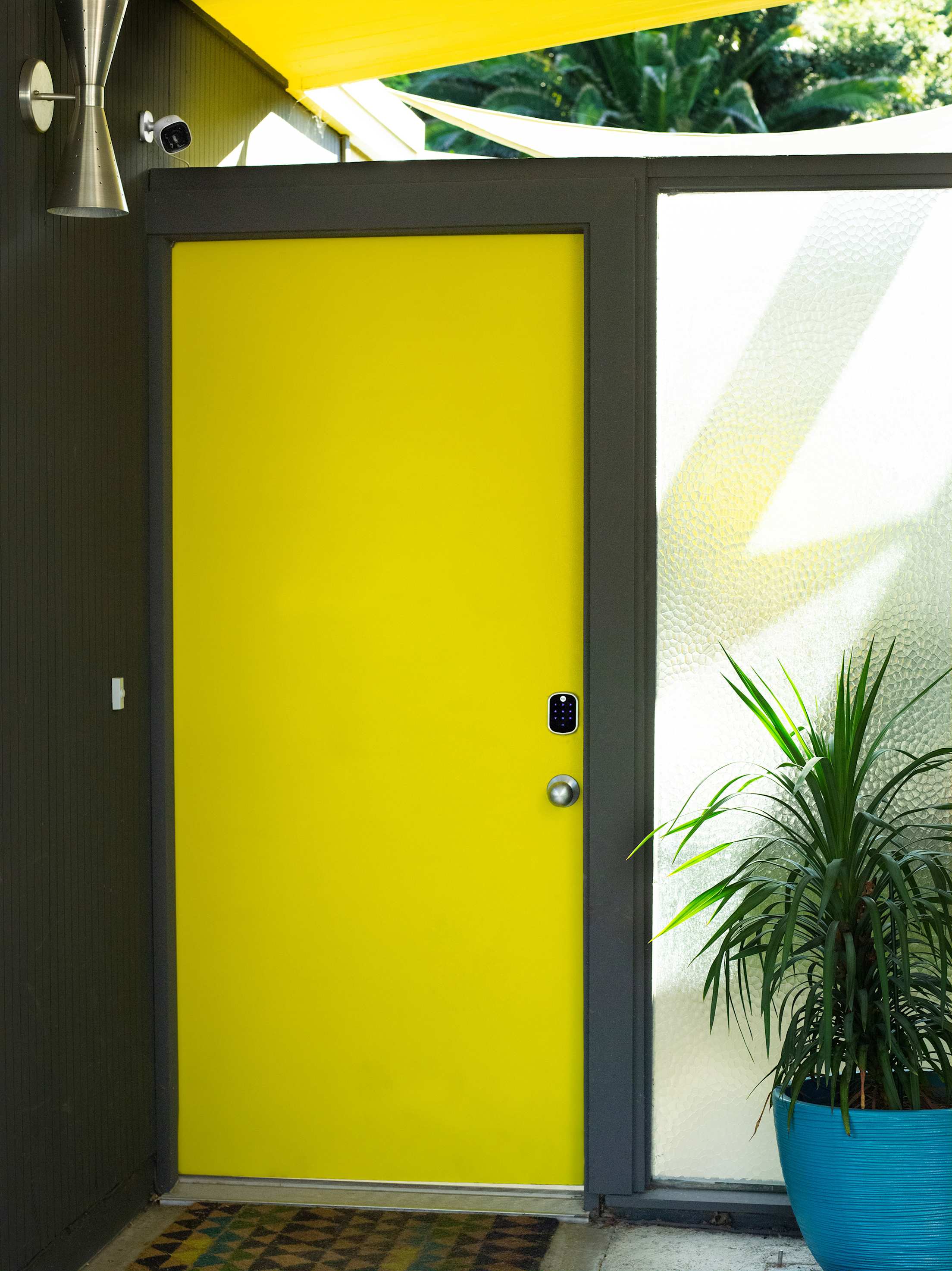 An outdoor security camera overlooks a porch and a smart lock on a yellow front door of a midcentury modern home.