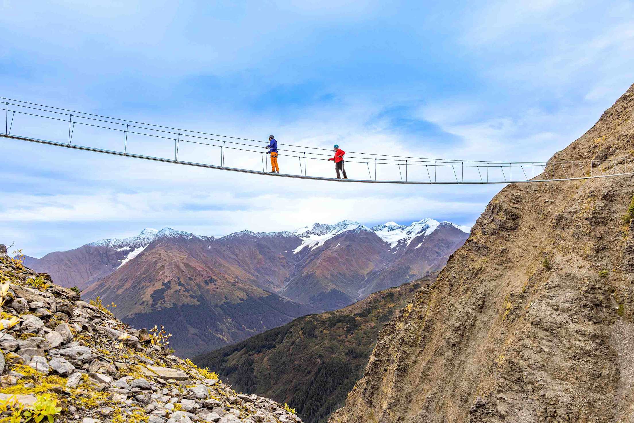 Two hikers navigate a cable bridge that runs between the peaks of Alyeska Mountain.