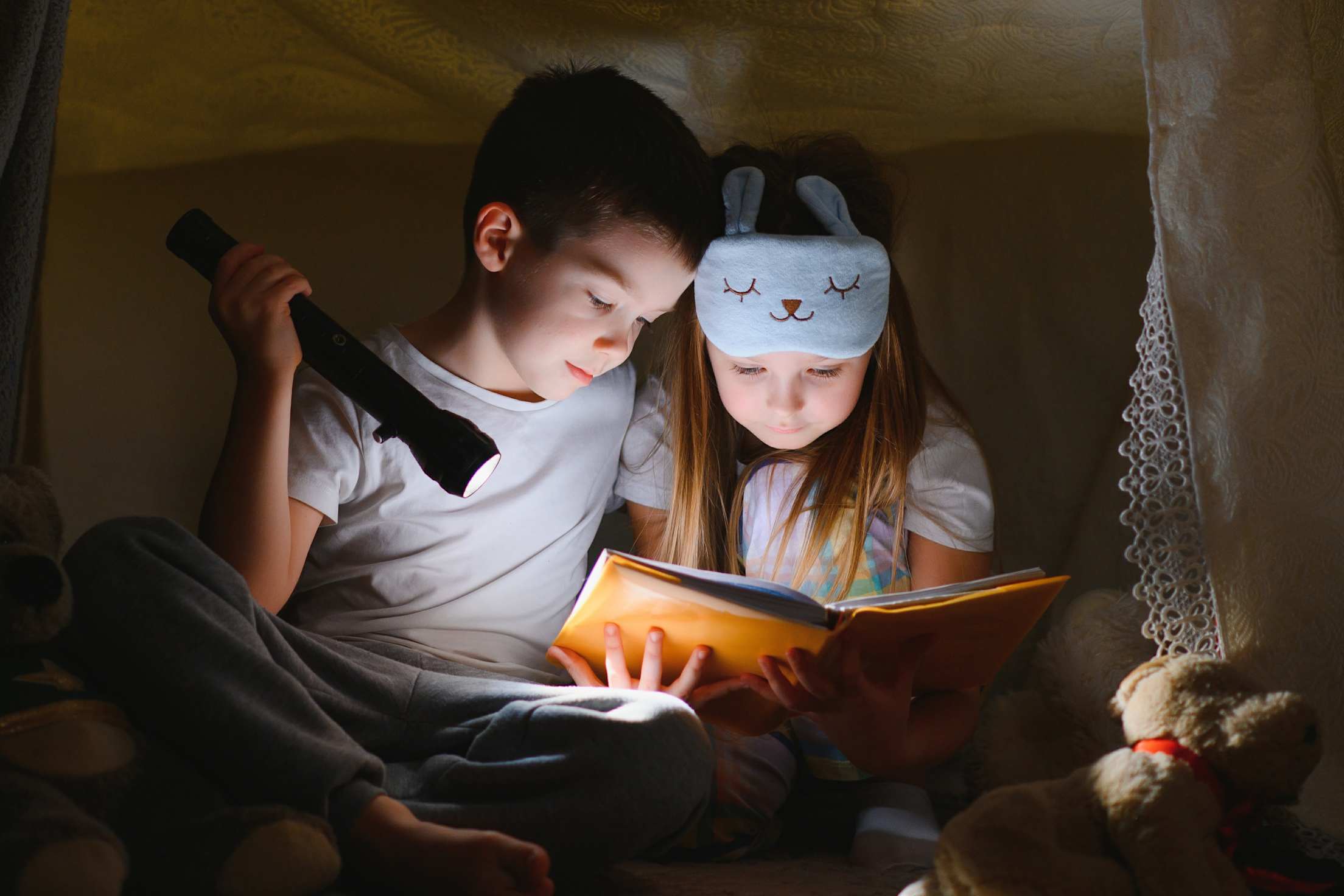 A brother and sister read a book by flashlight inside a blanket fort.