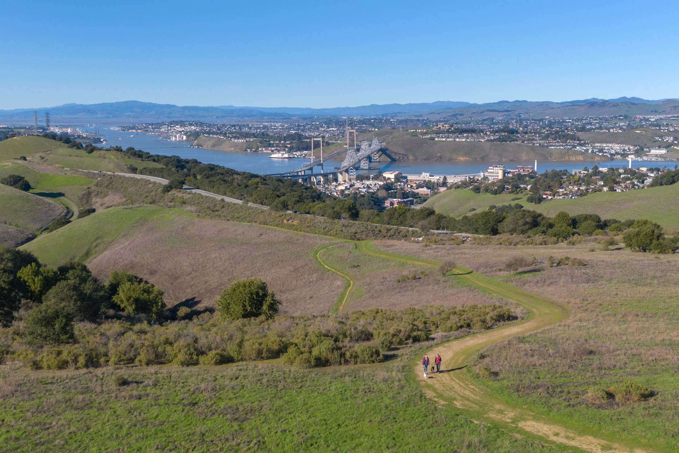 Two people and a dog hike on a trail in Crockett Hills Regional Park in Contra Costa County, California.
