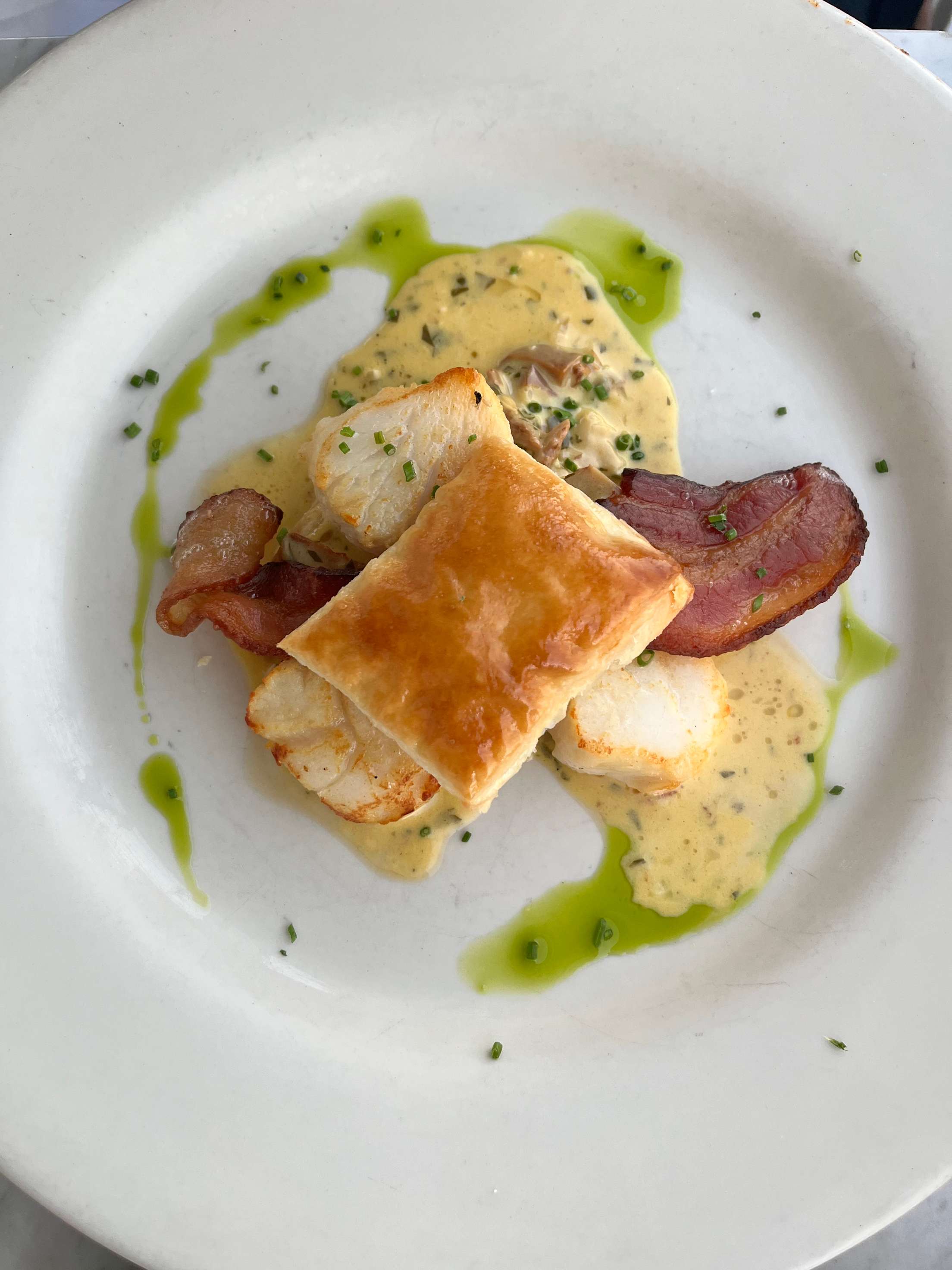 Pan-seared scallops with chanterelle duxelles, rasher of bacon, puff pastry, and chive oil.