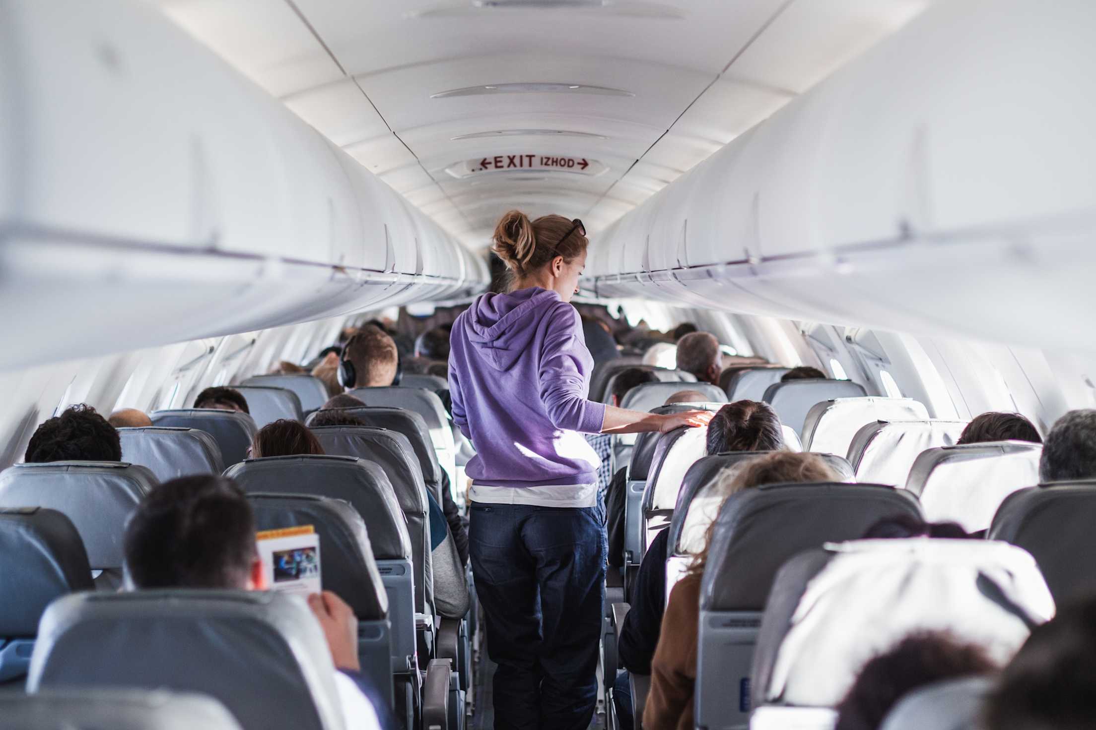 A woman in a purple sweatshirt walks up the aisle of a full airplane.