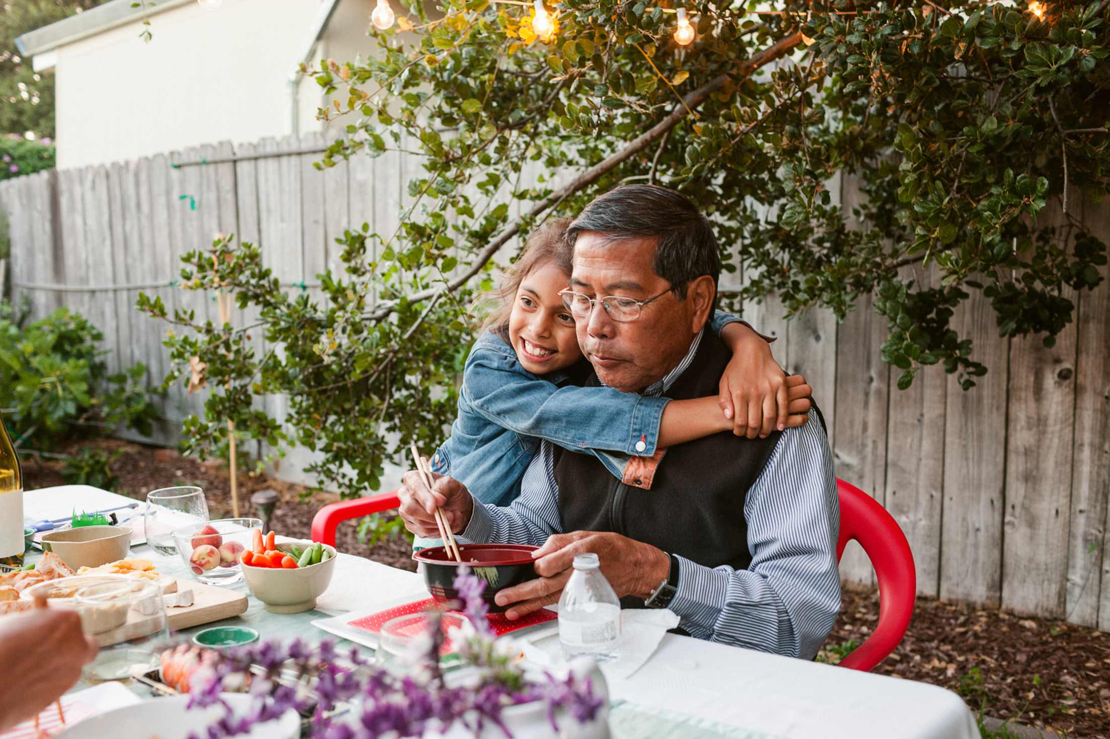 A granddaughter hugs her grandfather at an outdoor dining table.