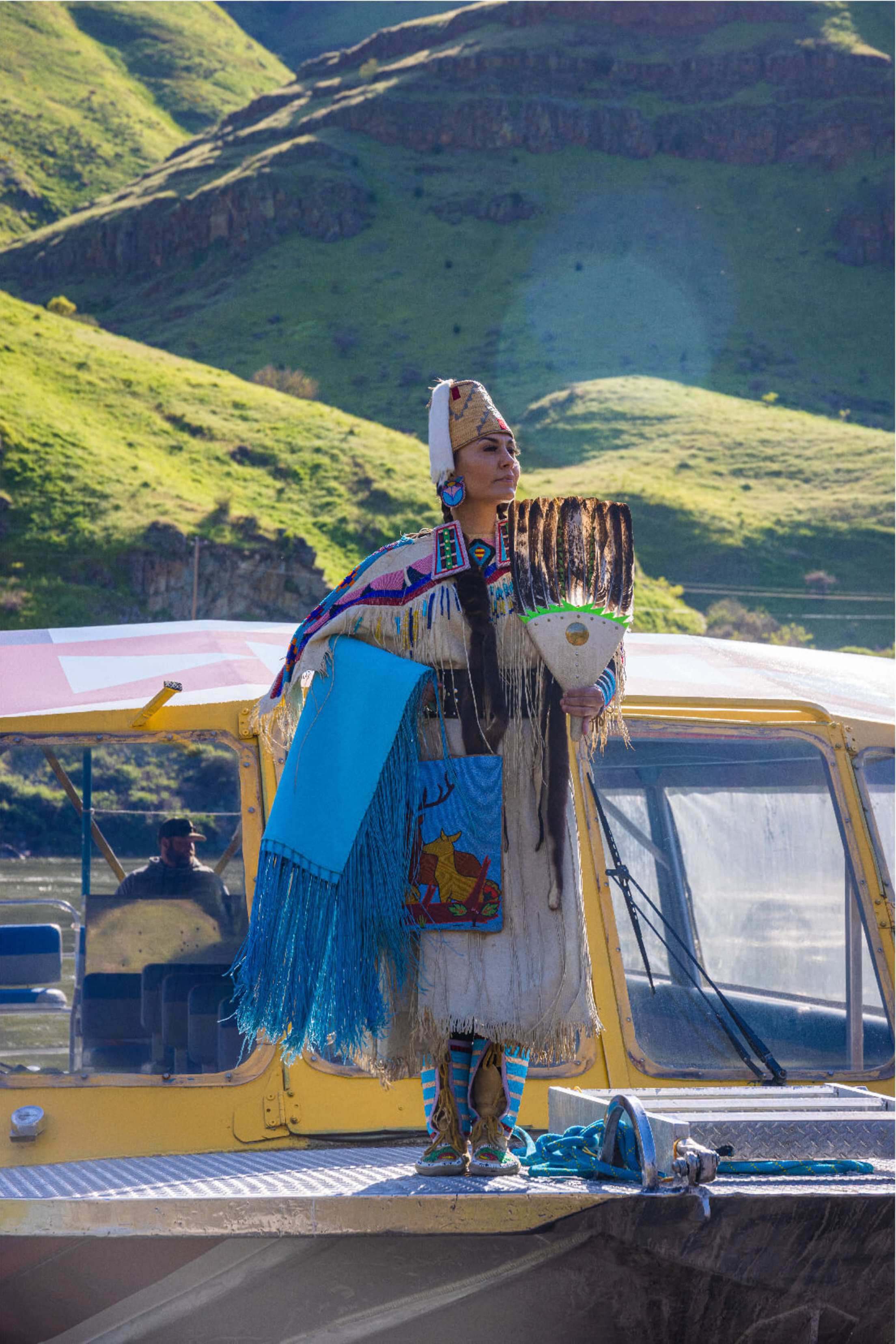 A Nimiipuu guide wearing colorful attire stands on a boat with green mountains behind her.