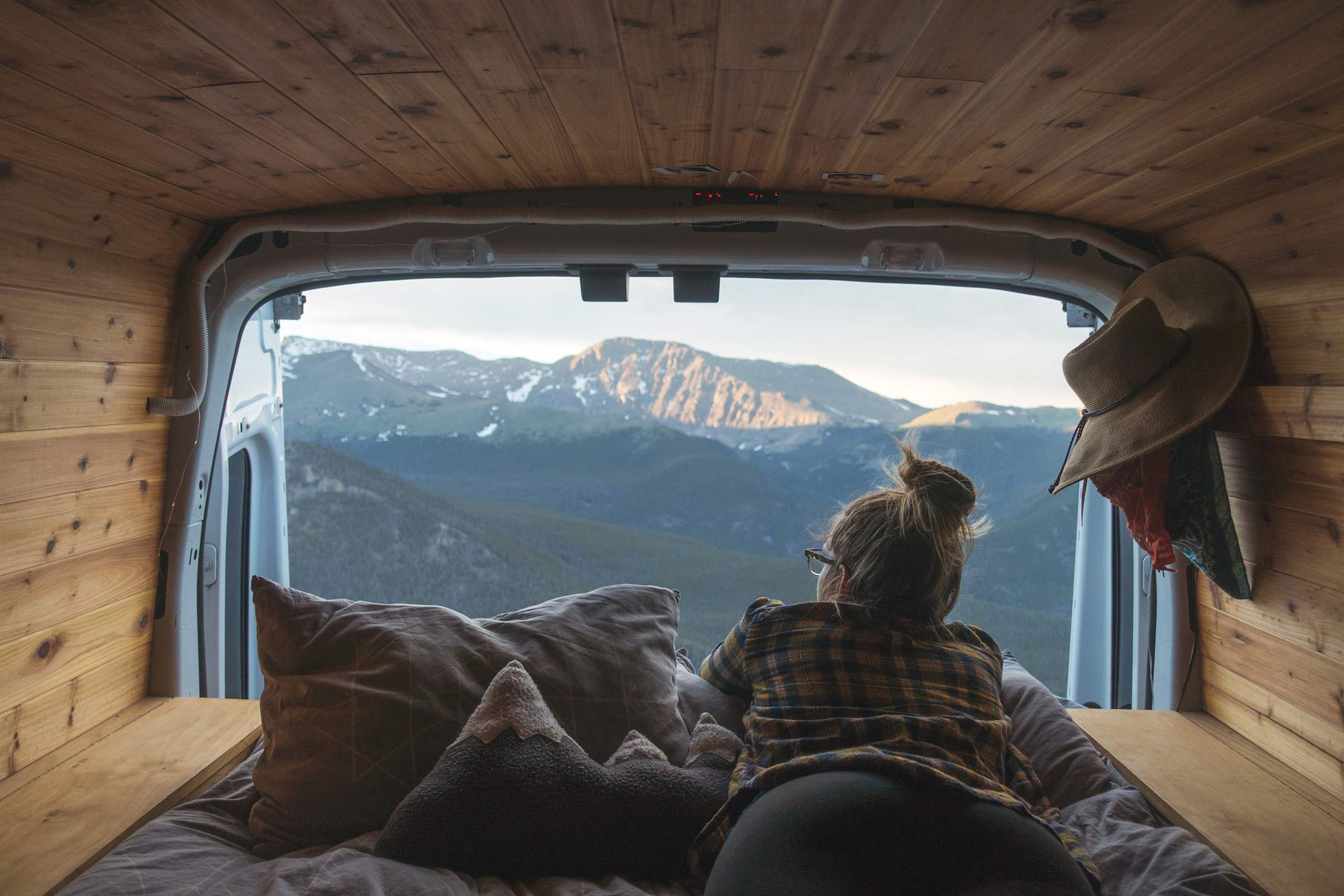 A woman looks out the back of a camper van at mountains out the back door