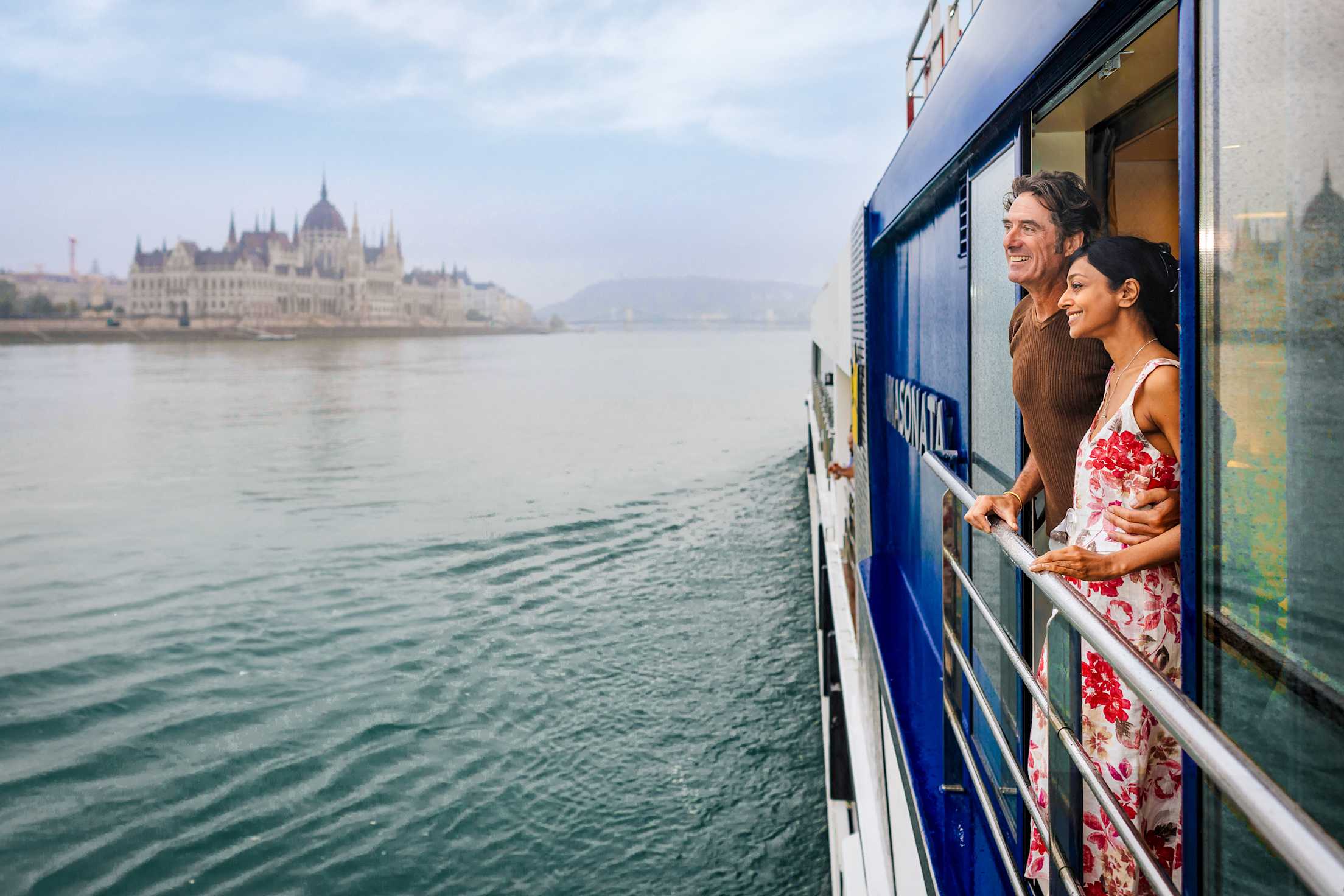 A man and woman standing on a river cruise ship balcony. The Budapest skyline is in the distance.
