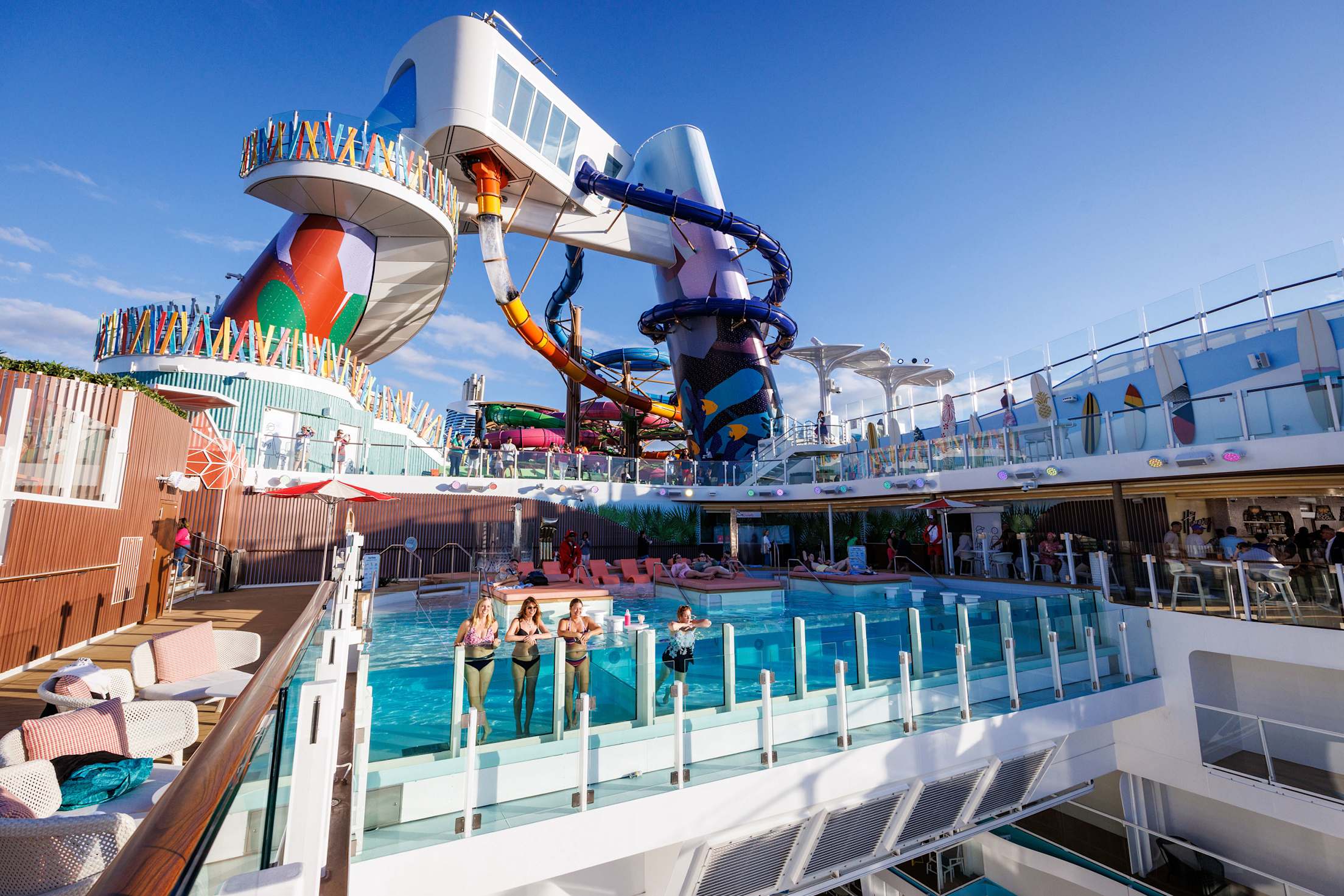 Adults in swimsuits stand along the see-through wall of a cruise ship pool in the daytime.