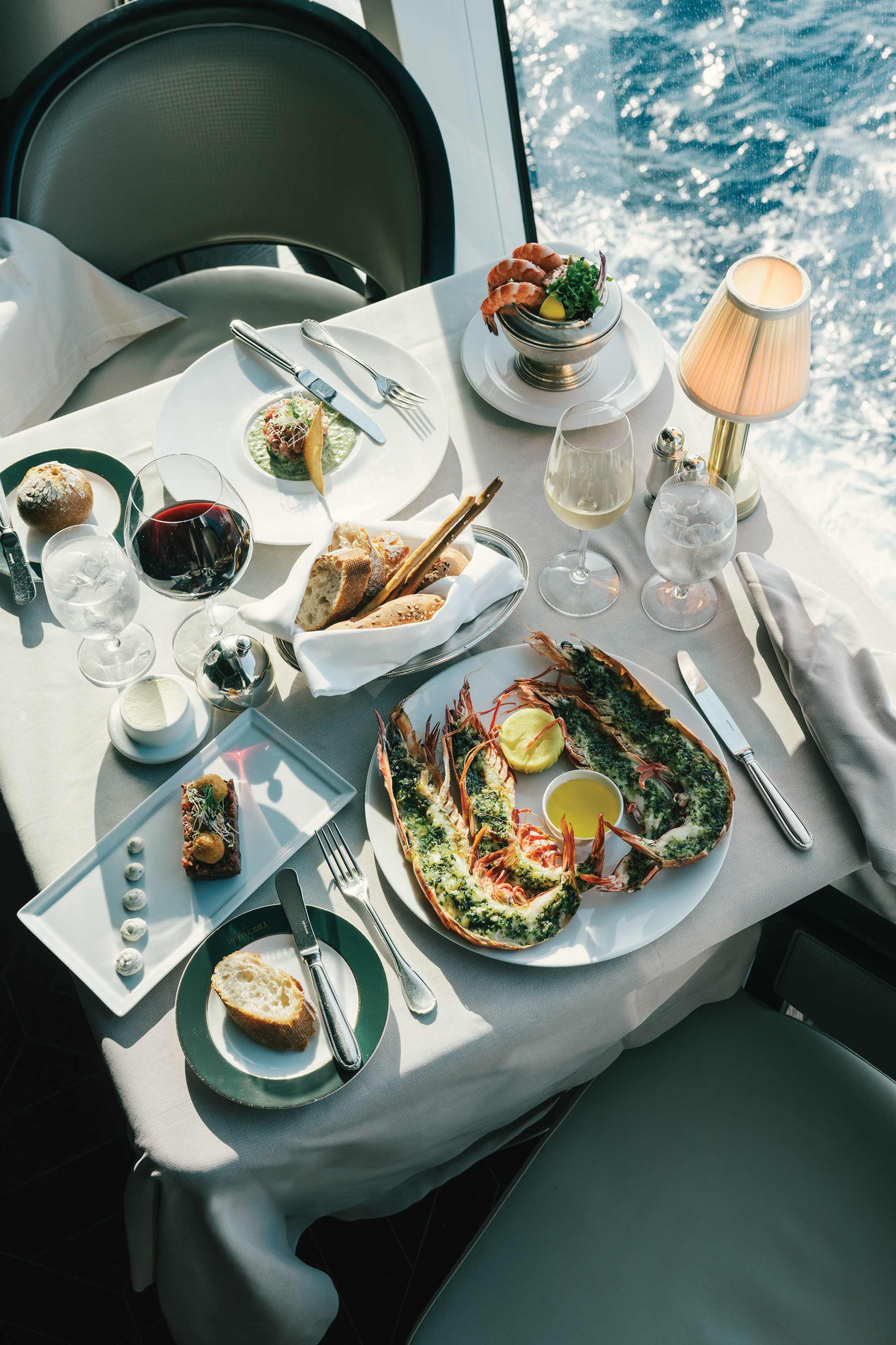 An ocean-view table for two with plated lobster, pasta, bread basket, and shrimp cocktail.